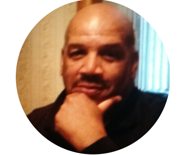 Headshot of Bennie Boyd. He is resting his head on his left hand and smiling warmly. Bennie is dark-skinned and bald, with a mustache. He is wearing a black top.