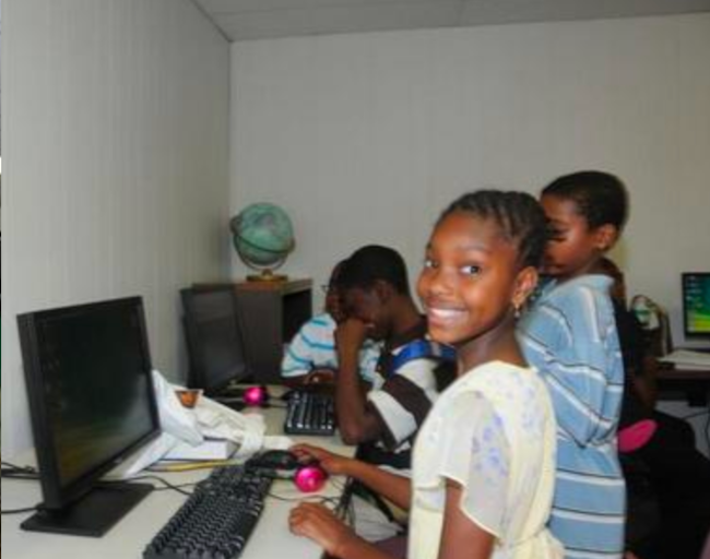 Children of color working in the GCLC computer lab. The girl in the foreground is looking up and smiling.