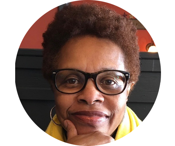 Headshot of J. Newsome. She is dark-skinned, has short brown hair, and is wearing black glasses. She is smiling and resting her chin on her hand.