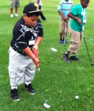 Children of color learning to play golf. They boy in the foreground is wearing a hat with a visor, a black-and-white long-sleeved jersey shirt with the sleeves hiked to the elbows, white baggy cargo shorts, and black sneakers. He is concentrating on properly holding a golf club. It is a sunny day. The grounds are green.